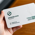 A close-up, eye-level shot of a professional individual's perfectly formed hands gently holding a modern business card. The card is crafted from a visibly thick, sustainable recycled paper, featuring a unique, subtle texture and a finely debossed, minimalist logo. The focus is on the tactile quality of the card and the natural pose of the hands, showcasing well-formed fingers and appropriate, modest accessories like a sleek watch. The background is a softly blurred, warm-toned professional office setting with natural light, emphasizing a sense of quality and meticulousness. fully clothed, appropriate attire, safe for work, appropriate content, professional, family-friendly, perfect anatomy, correct proportions, natural body proportions, proper finger count, professional photography, high detail.